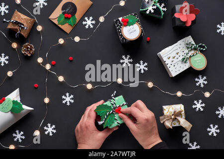 New Year or Christmas presents wrapped in various paper gift boxes with tags. Hands decorating box with holly. Flat lay, top view with light garland, Stock Photo