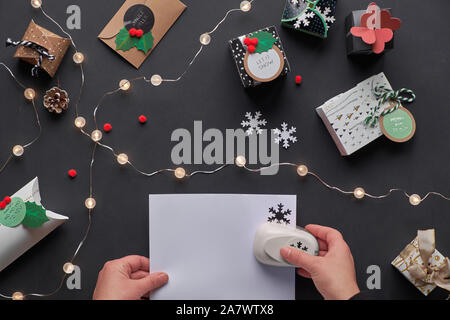 New Year or Christmas presents in various paper gift boxes with tags. Hands making paper snowflakes with hole puncher. Festive flat lay, top view with Stock Photo