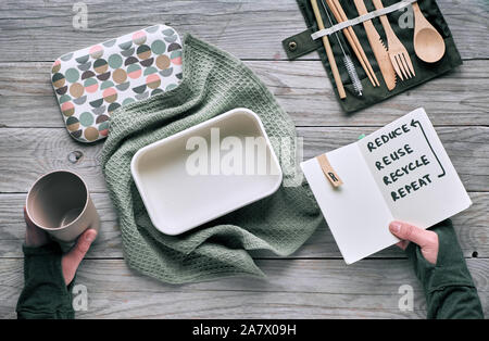 Flat lay, zero waste lunch - set of reusable wooden cutlery, lunch box in textile, reusable coffee cup. Sustainable lifestyle, hand holding notebook w Stock Photo