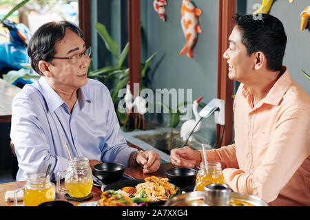Smiling mature man talking to his father when eating delicious Asian food in restaurant Stock Photo