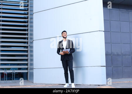 Young Arab businessman smiling and holds documents, stands near wall of business center. Stock Photo