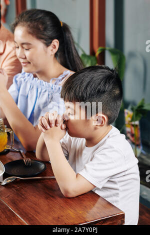 Adorable little Asian boy and his preteen sister praying before having meal at home Stock Photo