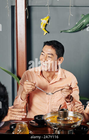 Portrait of happy middle-aged Vietnamese man eating noodles with spoon and chopsticks Stock Photo