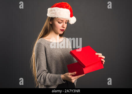 A model in a Santa Claus hat is disappointed in the gift. The girl opens the Christmas red gift and shows the emotion of surprise and disappointment. Stock Photo