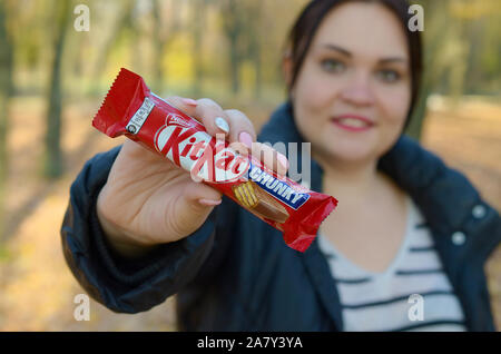 KHARKOV, UKRAINE - OCTOBER 21, 2019: A young caucasian brunette girl shows kit kat chocolate bars in red wrapping in autumn park. Kit Kat chocolate ma Stock Photo