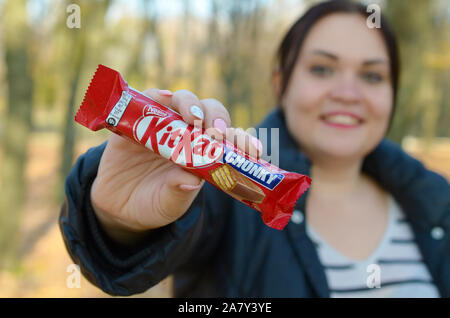 KHARKOV, UKRAINE - OCTOBER 21, 2019: A young caucasian brunette girl shows kit kat chocolate bars in red wrapping in autumn park. Kit Kat chocolate ma Stock Photo