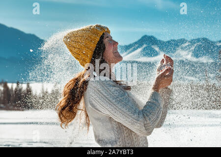 Happy beautiful girl plays with snow powder against forest and mountains