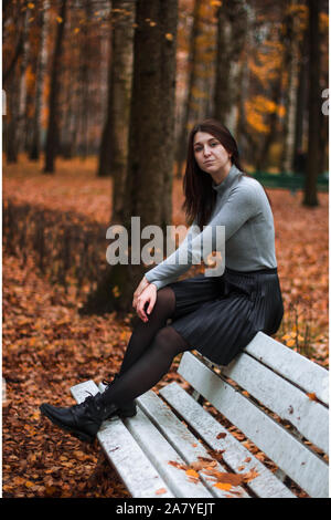Autumn outdoor portrait of beautiful young woman in autumn park, sitting on the back of a white bench and enjoying the fall landscape, wearing gray ba Stock Photo