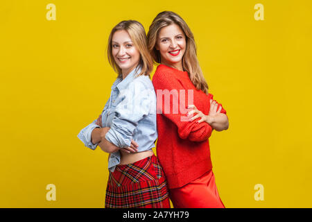 Best friends standing with their backs to each other and crossed hands. Portrait of two charming women in stylish casual clothes smiling at camera. in Stock Photo