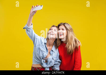 Female friends enjoying time. Portrait of two beautiful women in stylish clothes standing with toothy smile and taking selfie on phone, happy memories Stock Photo