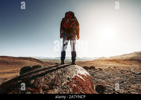 Hiker with backpack stands on big rock against mountains and sunset Stock Photo
