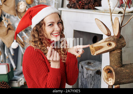 Laughing playful young woman in Santa hat pointing at wooden reindeer nose and looking at camera Stock Photo