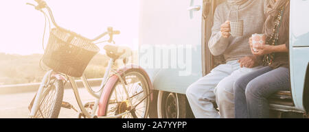 Unrecognizable adult senior couple sitting on an old vintage van together drinking tea or coffee - old bikes and sunlight in background - concept of l