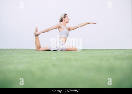 Beautiful bright image with caucasian girl doing pilates on the grass with white wall in background - healthy lifestyle for young millennial people en Stock Photo