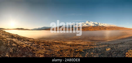 Amazing autumn panorama with the sun, mountains covered with snow and a golden forest, white fluffy fog over the lakes and the valley against the blue