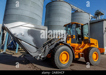 Yellow tractor infront of 3 Grain silos, in an industrial landscape, with a beautiful blue sky Stock Photo