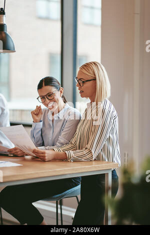 Two smiling young businesswomen reading paperwork together while sitting at a boardroom table during an office meeting Stock Photo