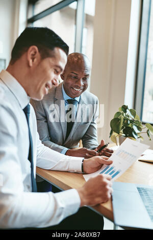 Two diverse businessmen smiling while sitting together at a desk in an office going over charts and graphs on paper Stock Photo