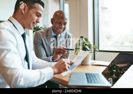 Two smiling diverse businessmen discussing charts and graphs on paper and using a laptop while sitting at a desk in an office Stock Photo