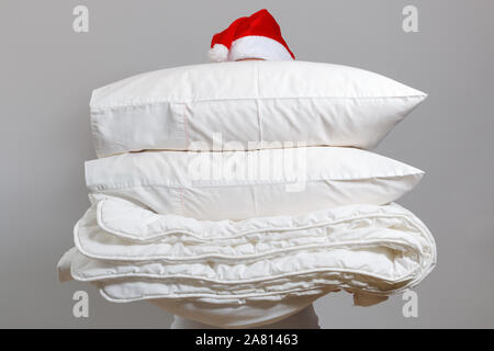 Woman in santa hat holding a warm duvet and feather pillows against a gray wall. Stack of bedding for sleeping. Household at Christmas. Stock Photo