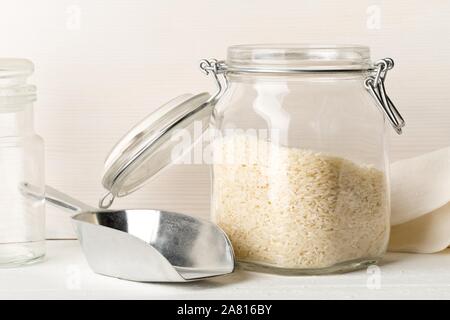 White uncooked, raw long grain rice in glass storage jar with metal scoop on white kitchen table background - selective focus Stock Photo