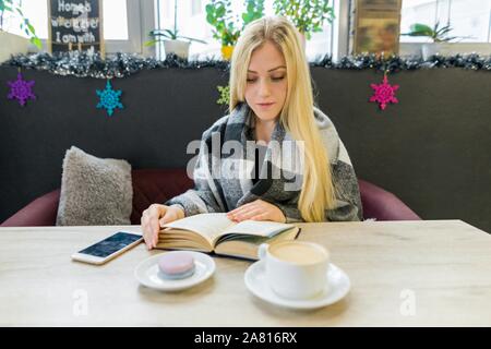 Young beautiful blonde girl sitting in coffee shop reading book. Winter season Stock Photo