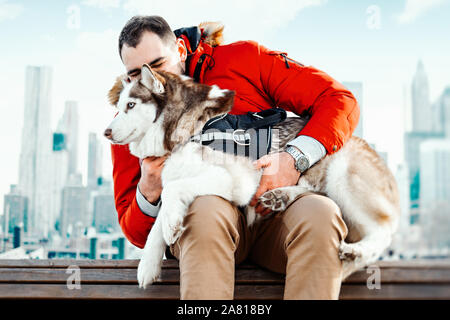 Man and Cute Siberian Husky dog sitting on bench on cityscape background on sunny winter day. Concept of animal wellbeing and owner care and love. Stock Photo
