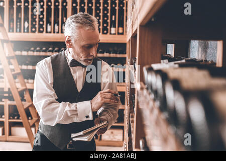 Sommelier Concept. Senior man standing checking wine collection notebook pensive Stock Photo