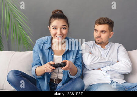 Relationnship Concept. Young couple man looking angry at woman playing video game concentrated sitting on sofa studio isolated on grey Stock Photo