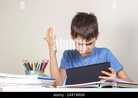 Sad confused child with tablet computer sitting at table with books notebooks Stock Photo