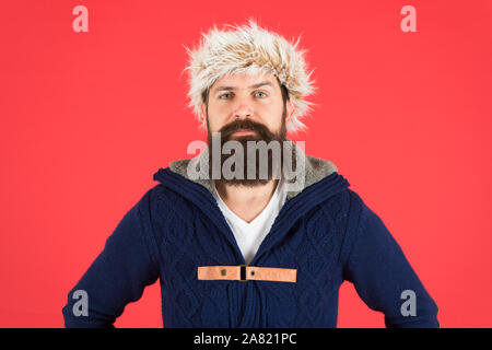 Winter menswear. Clothes design. Man bearded stand warm jumper and hat on red background. Winter season menswear. Hipster rustic style furry hat. Fashion menswear shop. Masculine clothes concept. Stock Photo