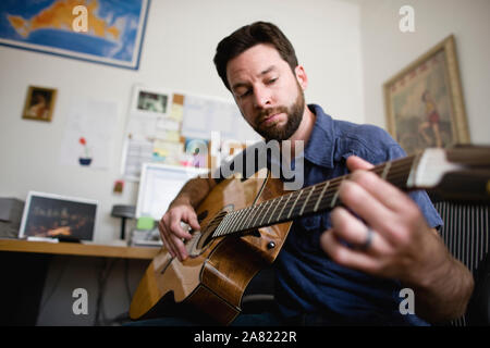 Bearded young man sitting playing an acoustic guitar inside a home  office. Stock Photo