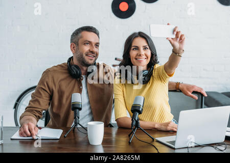 beautiful radio host sitting near handsome colleague and taking selfie with smartphone Stock Photo