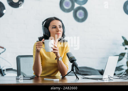 pensive radio host looking away while sitting at workplace and holding coffee cup Stock Photo