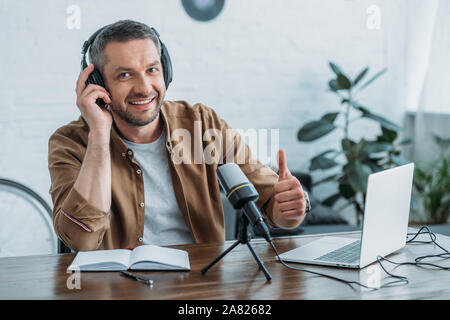 handsome radio host showing thumb up and smiling at camera while sitting at workplace Stock Photo