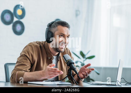 smiling radio host in headphones gesturing while speaking in microphone in broadcasting studio Stock Photo