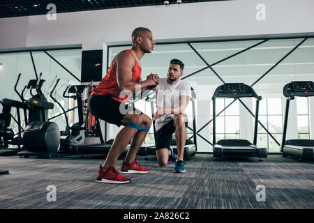 attentive trainer looking at young african american sportsman warming up in gym Stock Photo