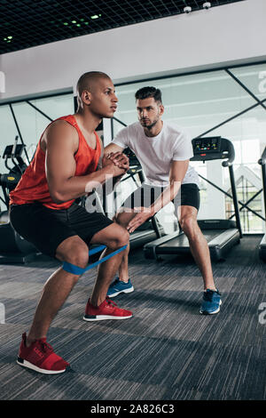 attentive trainer supporting african american sportsman warming up in gym Stock Photo