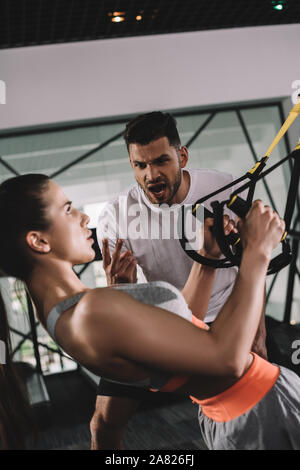excited trainer shouting while motivating young sportswoman pulling up on suspension trainer Stock Photo