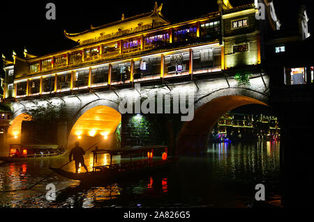 The nighttime silhouette of a man pushing a sampan boat, floats under the arches of the brightly lit Phoenix Hong Bridge in Fenghuang Ancient City in Stock Photo