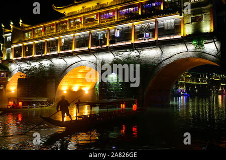 The nighttime silhouette of a man pushing a sampan boat, floats under the arches of the brightly lit Phoenix Hong Bridge in Fenghuang Ancient City in Stock Photo