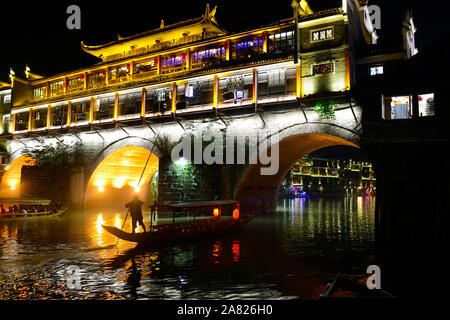 The nighttime silhouette of a man pushing a sampan boat, floats under the arches of the brightly lit Phoenix Hong Bridge in Fenghuang Ancient City in Stock Photo