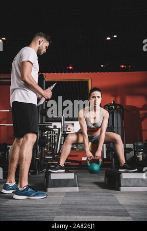 attentive trainer holding clipboard while standing near supervising young sportswoman lifting weight in gym Stock Photo