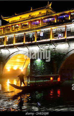 The nighttime silhouette of a man pushing a sampan boat, floats under the arches of the brightly lit Phoenix Hong Bridge in Fenghuang Ancient City in Stock Photo