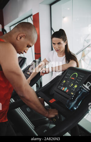 attentive trainer in fitness tracker looking at african american sportsman running on treadmill Stock Photo