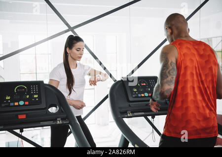 attentive trainer looking at fitness tracker while standing near african american sportsman running on treadmill Stock Photo