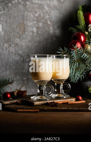 glasses with delicious eggnog drink on cutting board near decorated spruce branch on grey stone background Stock Photo