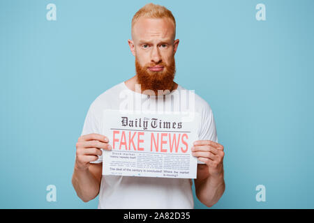 upset bearded man showing newspaper with fake news, isolated on blue Stock Photo