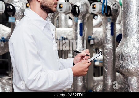 cropped view of bearded engineer in white coat holding clipboard and pen near air compressed system Stock Photo