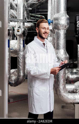 happy engineer in white coat holding pen and clipboard near air compressed system Stock Photo